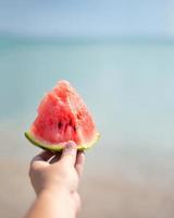 Watermelon slice in hand. Beach and sea. Ocean coast. Picnic on the coast. Vegetarian food in nature. photo
