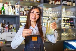 Beautiful female bartender is holding a shot glass with alcohol drink and a bottle in other hand, looking at camera and smiling while standing near the bar counter in cafe photo