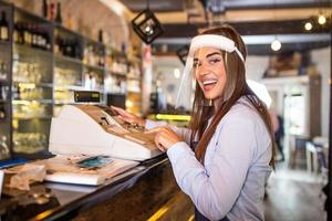 Waitress wearing an apron standing by a point of sale terminal and laughing while working in a restaurant. Beautiful woman wearing face shield during coronavirus pandemic standing by cash register photo