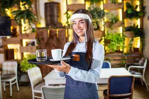 Female waitress wearing face shield, visor serves the coffee in restaurant during coronavirus pandemic representing new normal concept photo