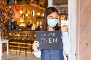 Happy female waitress with protective face mask holding open sign while standing at cafe or restaurant doorway, open again after lock down due to outbreak of coronavirus covid-19 photo