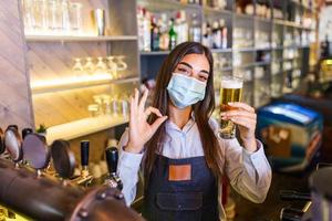 Bartender with protective face mask,covid-19 protection, serving a draft beer at the bar counter during coronavirus pandemic, showing OK sign,shelves full of bottles with alcohol on the background photo