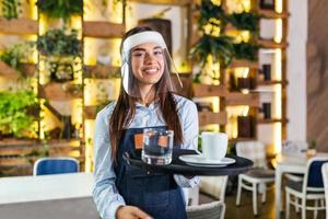 Female waitress wearing face shield, visor serves the coffee in restaurant during coronavirus pandemic. Holding tray with cup of coffee, working in cafeteria and serves the table photo
