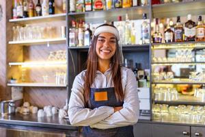 Portrait of a beautiful bartender standing at the counter smiling and looking at the camera while while wearing face shield due to covid-19, shelves full of bottles with alcohol on the background photo