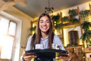 camarera feliz sosteniendo una bandeja con una taza de café, trabajando en la cafetería y sirviendo la mesa foto