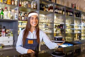 Beautiful female barista is holding a cup with hot coffee, wearing face shield while standing near the bar counter in cafe. New normal photo