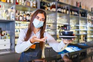 hermosa barista femenina está sosteniendo una taza con café caliente, mirando la cámara y usando una máscara protectora mientras está de pie cerca del mostrador del bar en el café foto