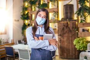 Happy female waitress using digital tablet while wearing protective face mask at the restaurant or cafe. Open again after lock down due to outbreak of coronavirus covid-19, New normal photo