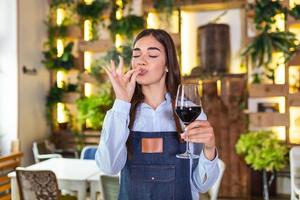 alegre y elegante camarera sosteniendo una copa de vino tinto mostrando un delicioso cartel, de pie en el restaurante. sommelier cata de vinos en el restaurante. foto