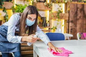 Waitress wearing protective face mask while disinfecting tables at restaurant or caffee for next customer. Corona virus and small business is open for work concept. photo