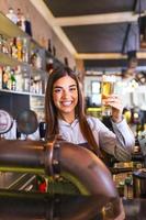 hermosa camarera sonriente que sirve una cerveza de barril en el mostrador del bar, mujer joven que sirve cerveza de barril. una camarera sonriente vertiendo cerveza fresca del grifo en el vaso en el pub. foto