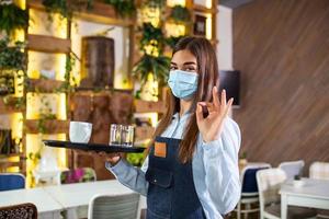 Happy waitress holding tray with cup of coffee, working in cafeteria and serves the table. Young woman wearing protective face mask during coronavirus pandemic photo