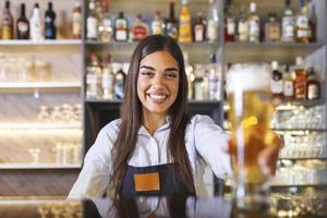 Beautiful smiling female Bartender serving a draft beer at the bar counter , shelves full of bottles with alcohol on the background photo