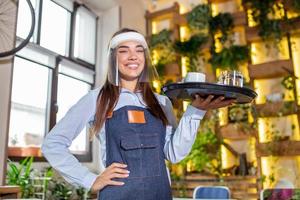 Female waitress wearing face shield, visor serves the coffee in restaurant during coronavirus pandemic representing new normal concept photo