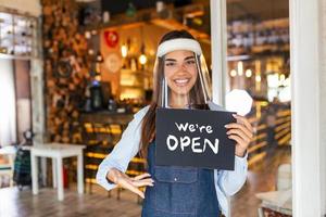 Small business owner smiling while holding the sign for the reopening of the place after the quarantine due to covid-19. Woman with face shield holding sign we are open, support local business. photo