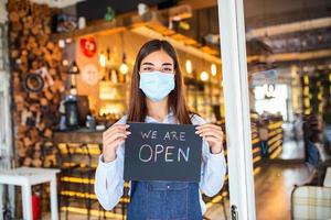 camarera feliz con mascarilla protectora sosteniendo un cartel abierto mientras está de pie en la puerta de un café o restaurante, abierta de nuevo después del cierre debido al brote de coronavirus covid-19 foto