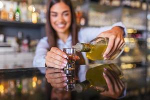 Beautiful female bartender is holding a shot glass with alcohol drink and a bottle in other hand, looking at camera and smiling while standing near the bar counter in cafe photo