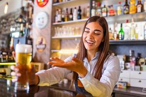 hermosa camarera sonriente que sirve una cerveza de barril en el mostrador del bar, estantes llenos de botellas con alcohol en el fondo foto