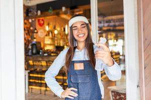 Waitress with face shield greeting customers in a restaurant or a caffe with bottle of alcohol to disinfect their hands before entering photo