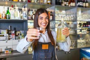 Beautiful female bartender is holding a shot glass with alcohol drink and a bottle in other hand, looking at camera and smiling while standing near the bar counter in cafe photo