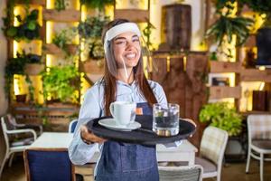 camarera con protector facial, la visera sirve el café en el restaurante durante la pandemia del coronavirus. sosteniendo una bandeja con una taza de café, trabajando en la cafetería y sirviendo la mesa foto