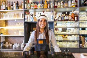 Portrait of a beautiful bartender standing at the counter smiling and looking at the camera while while wearing face shield due to covid-19, shelves full of bottles with alcohol on the background photo