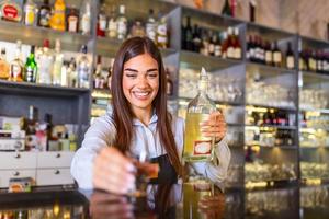 Beautiful female bartender with protective face maskholding shot glass with alcohol during coronavirus pandemic, shelves full of bottles with alcohol on the background photo