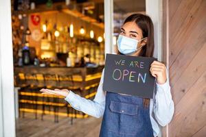Happy female waitress with protective face mask holding open sign while standing at cafe or restaurant doorway, open again after lock down due to outbreak of coronavirus covid-19 photo