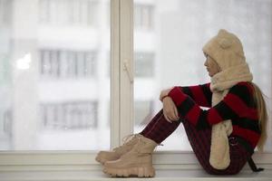 una joven adolescente pensativa se sienta en casa en el alféizar de la ventana y mira por la ventana, recuerda momentos tristes, se siente sola, piensa en problemas o errores. foto