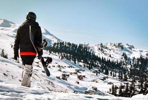 man tourist in ski suit with snowboard in his hands walks at snow-covered slope against the backdrop of beautiful mountain landscape photo