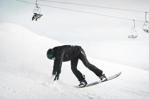 Extreme winter sports. Young professional male snowboarder rides performs tricks ride backwards pose for camera in ski resort bend snowboard. Black outfit snowboarder photo
