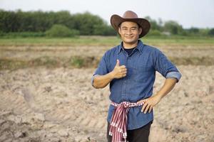 un agricultor asiático usa sombrero, camisa azul, pone la mano en la cadera y los pulgares hacia arriba, se siente seguro, se para en la tierra agrícola antes de cultivar. concepto, ocupación agrícola. granjero tailandés. foto