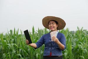 Asian female farmer wears hat, blue shirt, holds Thai banknote money and smart tablet at maize garden. Concept , Farmer gets agricultural supporting money via mobile app. Technology in agriculture.