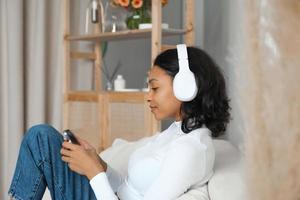Side view portrait of a happy african amercian girl listening music,podcast or lecture on sofa at home with phone in her hands. Online application and modern technologies concept. photo