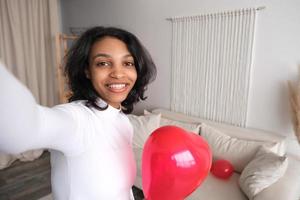 selfie retrato de feliz joven afroamericana con globo de corazón rojo. día de san valentín 14 de febrero o cumpleaños en apartamento moderno foto