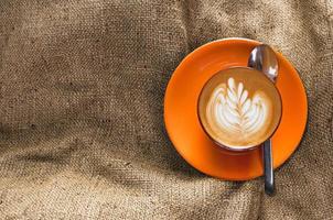 Top view hot latte in orange cup with floral pattern in foam on burlap background, brown sackcloth, vintage beige fabric texture photo