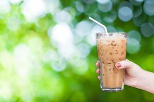 mano de mujer sosteniendo el café helado de vidrio sobre fondo verde de la naturaleza, café con leche helado foto