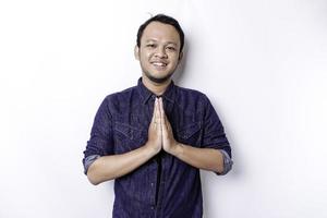 Smiling young Asian man wearing blue shirt, gesturing traditional greeting isolated over white background photo