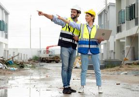 Engineer man and female architect wear safety helmets discuss housing development project at construction site using laptop computer. Contractor manager examining building estate infrastructure. photo