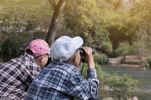 Asian boys using binoculars to watch birds on trees and fish in river in local national park during summer camp, idea for learning creatures and wildlife animals and insects outside the classroom. photo