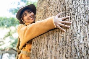 joven turista con un abrigo amarillo abrazando un árbol en el bosque del amor ecológico mirando las copas de los árboles joven asiática examinando un gran árbol ecológico foto