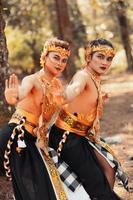 Two Balinese men dancing together in front of the tree while wearing a golden crown and golden necklaces in stripped clothes photo