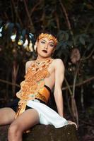 Sexy Asian man sitting on the big rock while wearing a golden costume and a golden crown photo