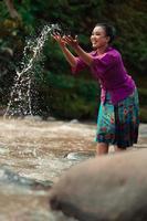 A beautiful Asian woman washing her hand and playing with the water while standing near the river in a traditional purple dress and green skirt photo