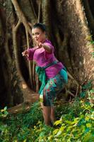 Sexy Asian woman dancing pose while wearing a beautiful purple dress and green belt on her body in front of the big brown tree photo