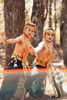 Two Balinese men dancing together in front of the tree while wearing a golden crown and golden necklaces in stripped clothes photo