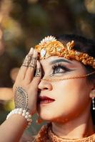 Asian woman covering her face with hands full of tattoos while wearing makeup and a golden headpiece photo