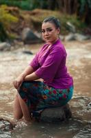 Beautiful Asian woman sitting on the rock near the river with a smiling face while wearing a purple dress photo