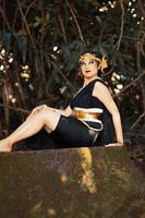 Indonesian woman sitting on the rock in a black dance costume while wearing a golden crown and golden necklace photo