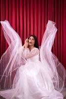 Elegance Asian woman wearing a wedding dress with flying fabric around her in front of the red curtain photo